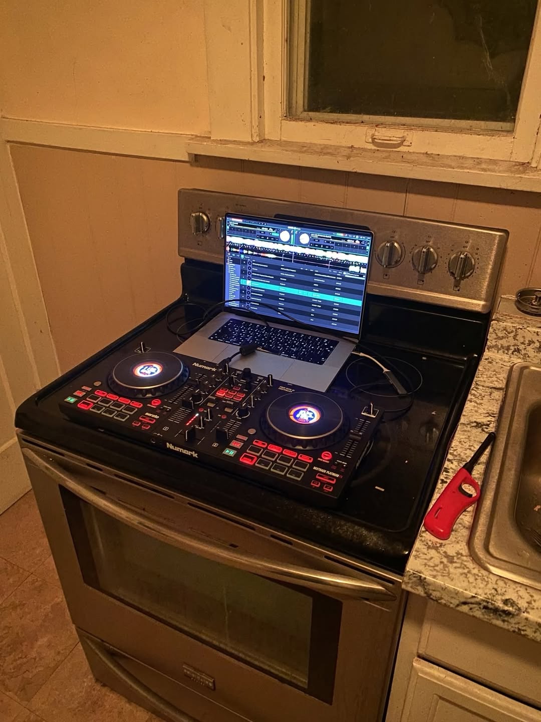 The image captures an absurd home setup where a Numark DJ controller and an open laptop running DJ mixing software are placed directly on the glass cooktop of a stainless steel electric kitchen oven. The scene is set in a residential kitchen: a granite countertop with a metal sink and a red utility lighter sits to the right of the stove, and a worn white-framed window is visible above the appliance. The comedic value comes from the unexpected, unsafe, and resource-poor choice to repurpose a cooking appliance as a stand for electronic music equipment, highlighting the relatable experience of hobbyists using whatever household surfaces are available when they lack dedicated studio furniture.