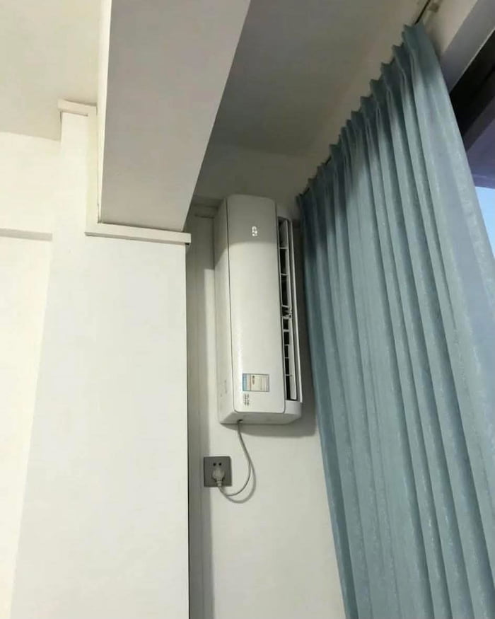 The image displays a wall-mounted split air conditioning unit installed in an extremely impractical, narrow gap between a protruding white structural partition on the left and a set of light blue pleated curtains on the right. The AC unit is positioned almost flush against the partition wall, meaning its front-facing air outlet vent is almost entirely blocked by the adjacent wall, preventing any cool air from circulating properly into the rest of the room. The unit is plugged into a wall socket below it, confirming it is connected to power. The core joke of the image is the implied question of whether this uselessly installed appliance can actually perform its intended cooling function.