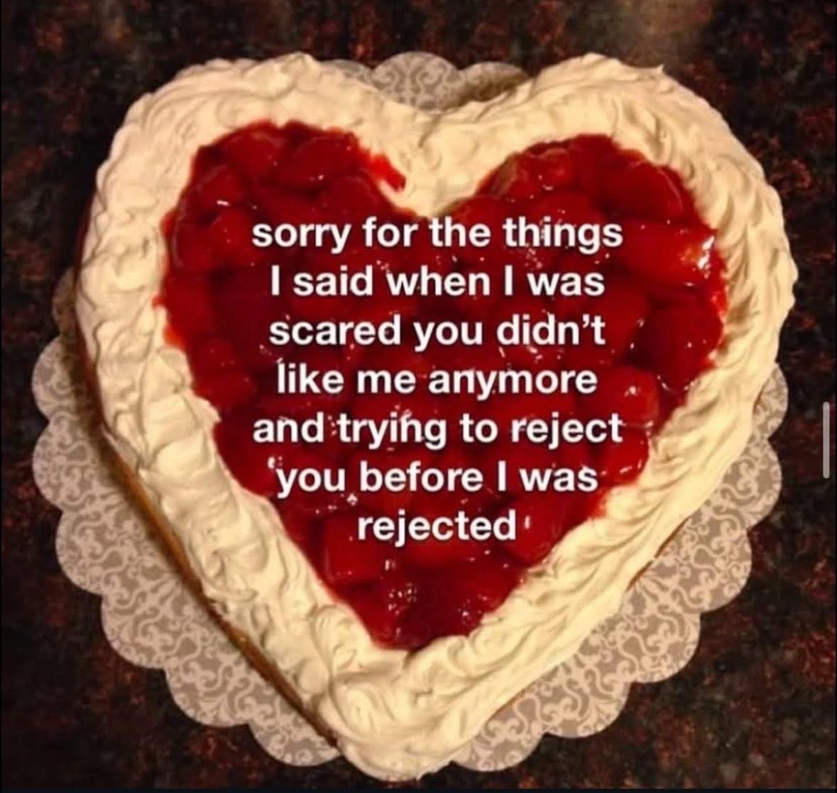 The image displays a heart-shaped cake with swirled white piped frosting along its border, and a center filled with glossy red fruit (likely strawberry or cherry) topping. White text is overlaid on the red fruit portion, stating an apology for hurtful things said when the speaker was afraid the other person no longer liked them, and had tried to reject the other person first to avoid being rejected themselves. The cake sits on a decorative scalloped paper doily on a dark patterned countertop.