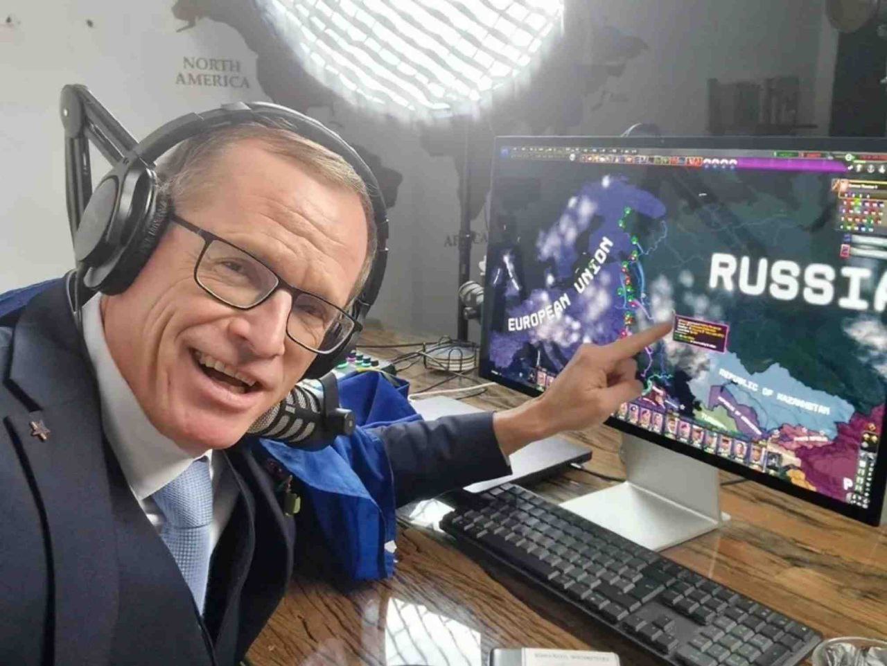 A man wearing headphones and glasses sits at a desk, pointing at a computer screen displaying a colorful political map with labels for 'EUROPEAN UNION' and 'RUSSIA'. Behind him is a wall with a world map visible. The man appears to be in a broadcast or analysis setting, wearing a suit and speaking into a microphone. The computer screen shows a detailed map with various colored regions and interface elements suggesting it might be from a strategy game or specialized mapping software.