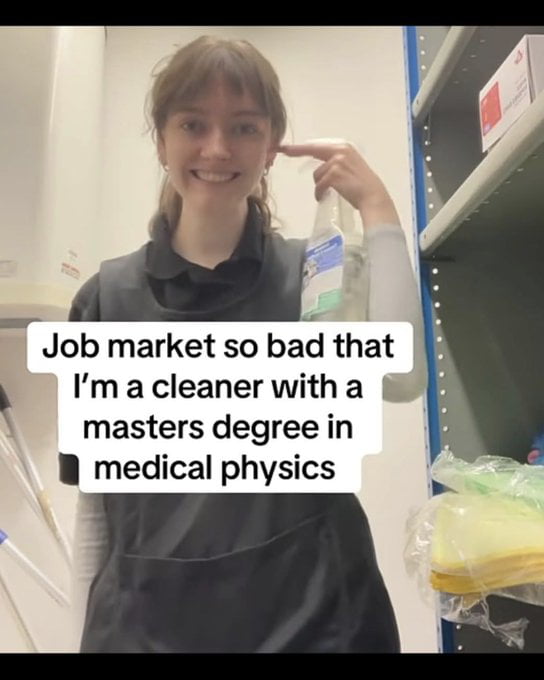 The image shows a young woman in a cleaning work apron holding a spray cleaning bottle, smiling and pointing to her head. Overlaid text on the image states that the job market is so bad that she is working as a cleaner despite holding a master's degree in medical physics. The meme highlights the widespread issue of underemployment for highly educated specialized graduates who are unable to find work in their field of study, forcing them to take low-skill jobs unrelated to their qualifications.