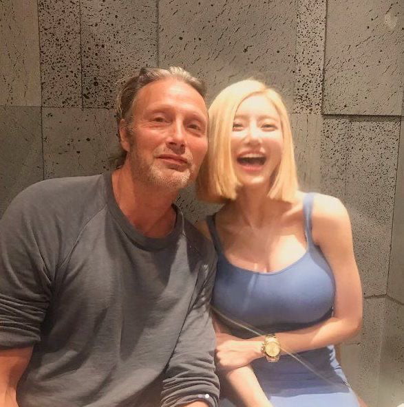 This post features a candid photo of middle-aged actor Mads Mikkelsen posing beside significantly younger Korean DJ Soda, who is laughing happily at the camera. The post is paired with the question "Guys should we dating someone younger?", using the clear large age gap between the two people in the photograph as context to prompt discussion about age gap romantic relationships. No additional text or edits are overlaid on the original photo itself.