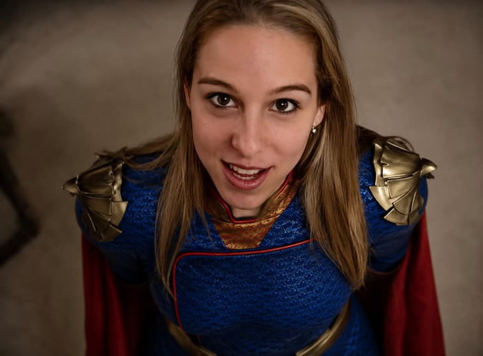 The image is a high-angle shot looking down at a woman wearing a genderbent Homelander cosplay costume, consisting of a blue textured suit with gold eagle-shaped shoulder armor, gold and red trim at the neckline, and a red cape. She has an open-mouthed, shocked, almost desperate pleading expression as she looks upward toward the camera. The visual is used as a joke to visualize the normally arrogant, tyrannical superhero Homelander from the series *The Boys* immediately becoming helpless and pathetic the moment he loses all of his superpowers.