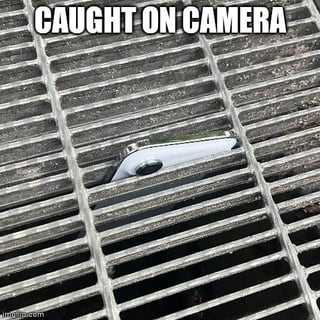 The meme displays a photograph of a single white wireless AirPod wedged between the metal grates of a public street storm drain, peeking out from under the grates as if it is hiding or peeking up at the person taking the photo. Large, bold white text reading 'CAUGHT ON CAMERA' is overlaid at the top of the image, humorously framing the common frustrating experience of losing small wireless earbuds in inaccessible places as if the AirPod is a suspicious or noteworthy subject being filmed for a surveillance or news segment.