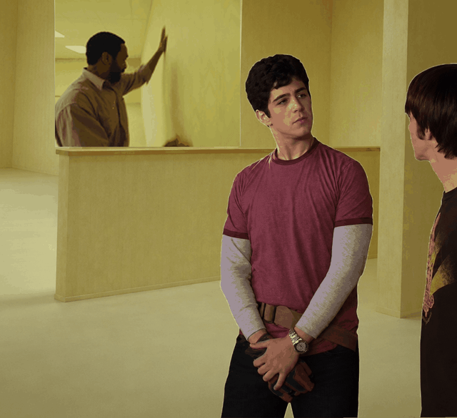 The GIF depicts a scene set in a pale yellow institutional hallway. In the foreground, two young men are facing each other in conversation, one of whom is the character Jimmy Brooks played by Aubrey 'Drake' Graham. In the background, behind a low half-wall partition, a man repeatedly pats and slaps at a completely blank, smooth wall that has no door opening, miming the action of searching for or trying to open a door that does not exist. The joke ties to the famous line 'Where's the door hole, Drake?' from the source show, referencing an infamous poorly made set piece that lacked a cutout for a door, leading to the humorous line.