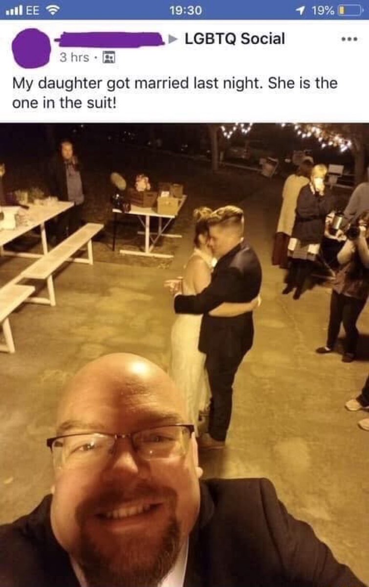 This image is a screenshot of a social media post shared in an LGBTQ Social group. The text of the post states 'My daughter got married last night. She is the one in the suit!'. The accompanying photo is a close-up selfie of a bald, glasses-wearing man in a suit smiling directly at the camera. In the background of the photo, a lesbian couple is dancing at their wedding: one wears a traditional wedding dress, while the other wears a formal suit. The comedic misdirection of the meme comes from the fact that viewers first incorrectly assume the man in the foreground taking the selfie is the daughter referenced in the post caption, before realizing the daughter is actually the suited person dancing with her spouse in the background.