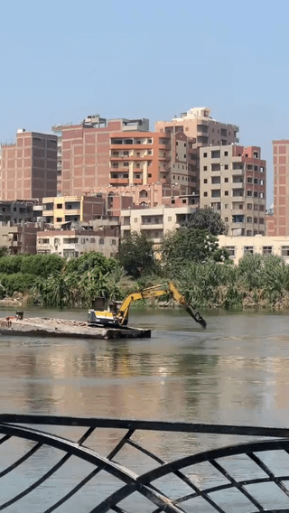 The image is captured from a bridge, with a black metal bridge railing visible in the immediate foreground. The middle ground features a wide, murky river (the Nile, consistent with the Egyptian setting), where a yellow industrial excavator is positioned on a small, low floating concrete platform in the middle of the water. The excavator's articulated arm is extended down into the river, appearing to dig or perform construction work directly in the water. Along the far riverbank, there is dense green vegetation, behind which stands a cluster of mid-rise, mostly red-brick and beige residential apartment buildings typical of urban Egyptian riverfront areas, under a clear pale blue sky. The humor comes from the absurdly unexpected sight of heavy construction equipment operating in the middle of a major river, which the title frames as an unremarkable, 'normal' everyday occurrence in Egypt.