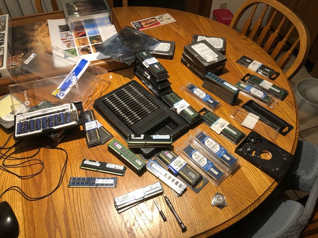 A photograph of a cluttered wooden table filled with numerous computer hardware components, primarily various RAM (Random Access Memory) modules of different sizes and form factors, some still in original packaging and others loose. Additionally, there are a few hard disk drives, small tech parts, and packaging materials scattered across the table. The scene humorously suggests the owner has a large stockpile of RAM, playing on the context of a current RAM shortage to joke about cashing in the collection like a retirement portfolio.