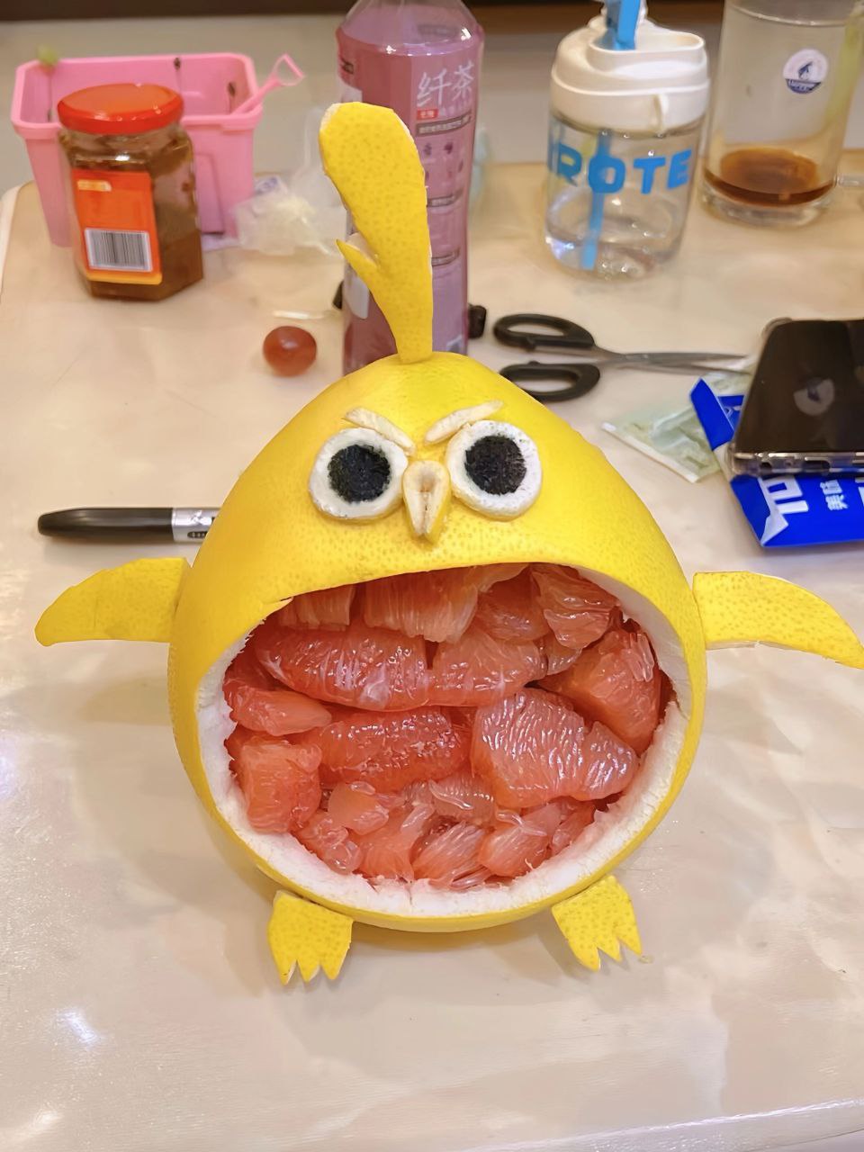 The image showcases a creative hand-carved pomelo (grapefruit) sculpture designed to resemble a playful bird or owl. The yellow pomelo peel is skillfully shaped with a rounded body, small wings, and a pointed crest on top. The creature's face features large, expressive eyes made from contrasting materials (likely black and white paper or fabric), a tiny triangular beak, and an open 'mouth' revealing vibrant pink pomelo segments arranged inside, mimicking teeth or a tongue. The sculpture is placed on a light-colored table surrounded by various everyday items: a pink container, a jar with an orange label, a purple bottle with Chinese characters '纤茶' (Qian Cha, a tea brand), a clear water bottle labeled 'ROTE', black-handled scissors, a silver pen, a smartphone, blue packaging, and a single red grape. The overall scene conveys a sense of casual creativity and DIY artistry, highlighting the maker's ingenuity in transforming a common fruit into an adorable decorative piece.