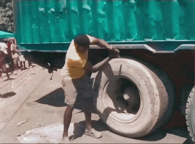 This image is a still from a perfectly timed short video. It shows a Black man in a yellow t-shirt, beige shorts and slip-on shoes, bending over to work on a large truck tire with a crowbar. The shadow cast by his arm holding the crowbar aligns perfectly with his lower body, creating a funny optical illusion that makes it look like he is inserting his own leg into the center of the truck tire rim, as if he is attempting to remove his leg rather than repair the tire. The scene takes place outdoors on a paved road, with a large green truck and a group of bystanders visible in the background, and the image has a slightly grainy, vintage filter effect.
