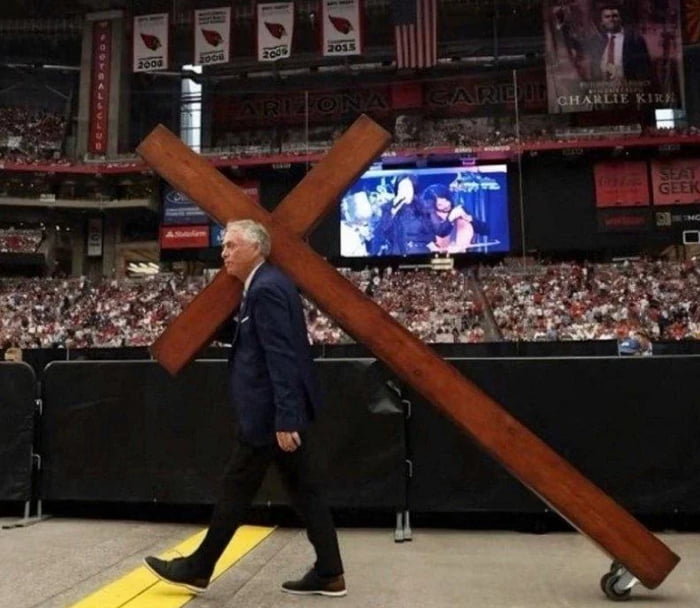 An edited image depicting an older man in a dark suit carrying a large wooden cross with wheels attached to its base. The scene is set in a packed stadium identified as the Arizona Cardinals' venue, with visible banners reading 'ARIZONA CARDINALS' and years like '2008' and '2015'. An American flag hangs from the rafters, and a large video screen in the background displays indistinct figures. The cross, modified with wheels to ease transportation, satirizes the idea of symbolic sacrifice without actual effort.