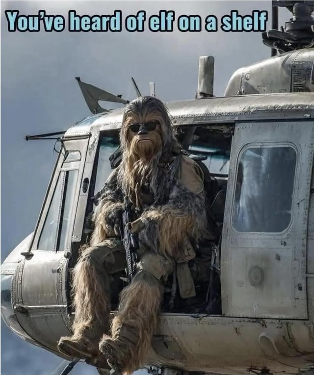 A humorous meme image featuring Chewbacca, the Wookiee character from Star Wars, seated in the open door of a military helicopter. He is dressed in his typical furry costume, wearing sunglasses, and holding an assault rifle. The top of the image contains the text 'You've heard of elf on a shelf,' which is a playful pun referencing the popular Christmas tradition 'Elf on a Shelf,' replacing 'elf' with Chewbacca and 'shelf' with the helicopter (a 'chopper'). The image combines elements of Star Wars with a military helicopter setting, creating a comedic contrast to the innocent holiday tradition.