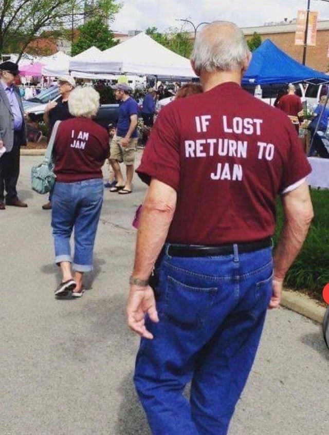 A photograph capturing an outdoor market scene with two individuals walking away from the camera. The foreground figure wears a maroon shirt with bold white text on the back: 'IF LOST RETURN TO JAN'. Behind them, another person (presumed to be their partner) sports a matching maroon shirt with the text 'I AM JAM' printed on the back. The market environment includes multiple white and blue tents, other attendees engaging in casual activities, parked cars, and greenery, creating a lively, community-focused atmosphere. The shirts feature a playful pun, as 'Jam' phonetically mirrors 'Jan', suggesting a lighthearted, affectionate dynamic between the couple.