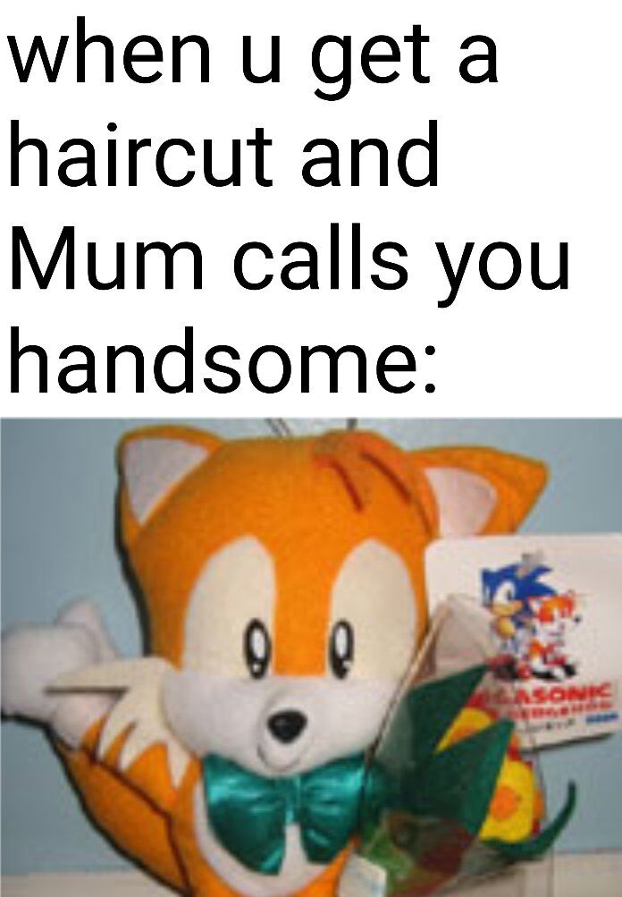 The image consists of two main parts: a top text section and a lower photograph. The top text reads, 'when u get a haircut and Mum calls you handsome:' in bold, black font on a white background, setting up a relatable scenario of positive parental feedback after a haircut. Below the text is a photograph of an orange fox plush toy with white facial markings and a green bow tie, identified as Tails from the Sonic the Hedgehog franchise. The plush has a happy, content expression with large, round eyes and is positioned as if waving one hand while holding a small Sonic-themed item (likely a card or merchandise featuring Sonic and Tails). The overall tone is wholesome and endearing, using the cute plush toy to visually represent the feeling of joy from being called handsome by one's mother.