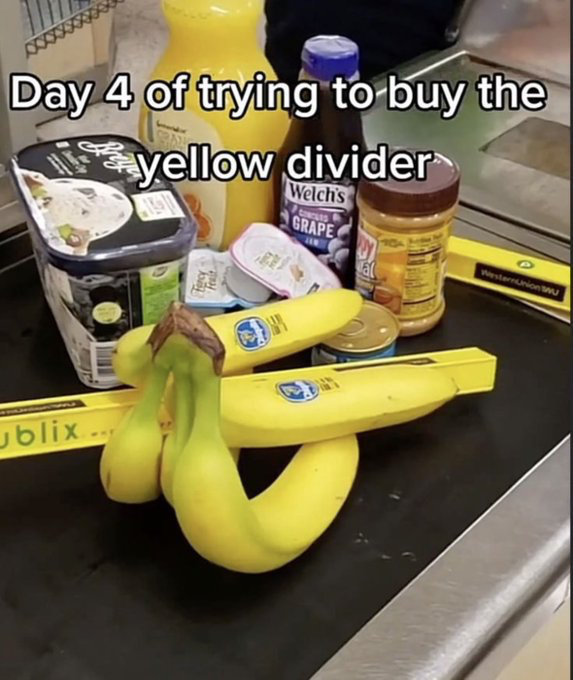 This meme shows a grocery store checkout conveyor belt with standard grocery items, official yellow Publix checkout dividers, and a bunch of bright yellow bananas positioned to look exactly like another checkout divider. The overlaid text reads 'Day 4 of trying to buy the yellow divider', creating a funny visual gag: the poster playfully pretends they believe the banana bunch is a store-owned checkout divider that they are attempting to purchase, implying cashiers have repeatedly told them it is not for sale over multiple trips. The joke relies on the visual similarity between the long, yellow bananas and the equally long, yellow store dividers.