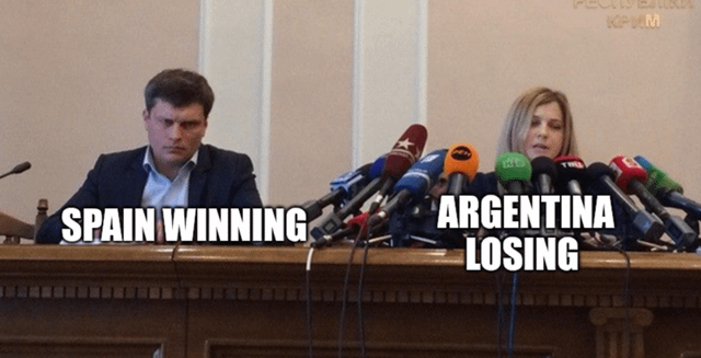 This meme uses a press conference photograph to satirize media coverage trends around FIFA football matches. A man on the left side of the table has almost no microphones directed at him, labeled 'SPAIN WINNING', while a woman on the right is completely surrounded by dozens of press microphones, labeled 'ARGENTINA LOSING'. The joke highlights that audiences and sports media pay vastly more attention to Argentina losing or underperforming in matches than they do to Spain winning their matches, a recurring trend in recent FIFA tournament discourse.