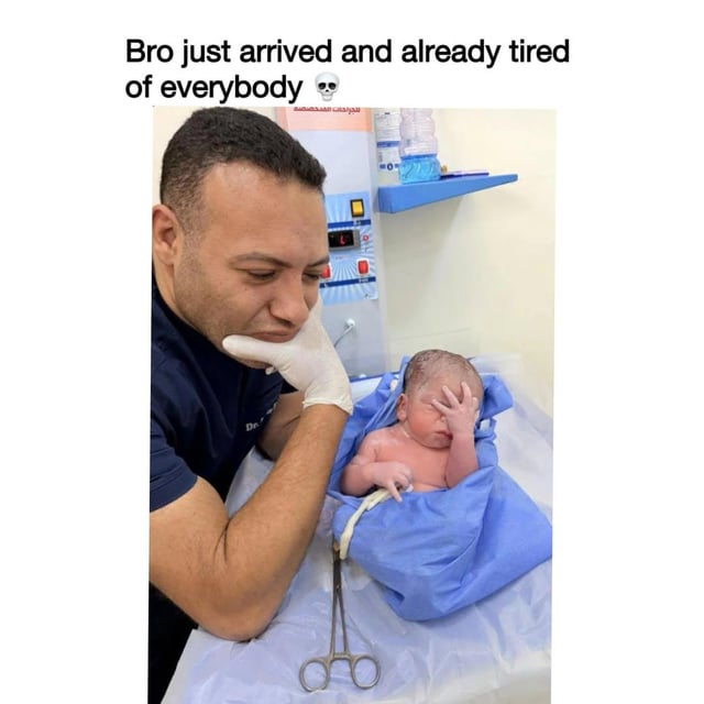 This meme features a candid photograph taken in a hospital setting. A male medical professional wearing dark scrubs and sterile white gloves leans on a medical table, resting his chin on his gloved hand as he looks at a newborn baby. The newborn is wrapped in a blue medical cloth, lying on the table next to a pair of surgical scissors. The baby is holding one hand up to its forehead in a classic facepalm gesture, appearing exhausted, annoyed, or unimpressed. Text placed above the photo reads 'Bro just arrived and already tired of everybody' paired with a skull emoji, humorously framing the baby's pose as the baby already being fed up with people right after being born, a relatable joke for many people who feel similarly tired of social interactions.