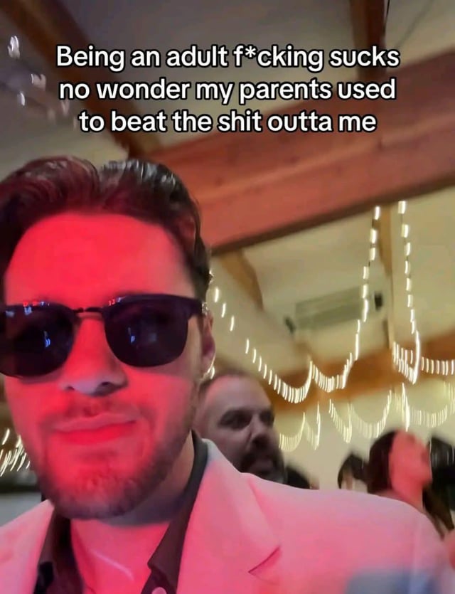 This is a single-panel selfie meme showing a man wearing dark sunglasses and a pale pink blazer, with his face lit by red ambient party lighting. He has a deadpan, neutral expression while taking a selfie at an indoor event, with string lights and other party guests visible out of focus in the background. Overlaid white text at the top of the image reads: 'Being an adult f*cking sucks no wonder my parents used to beat the shit outta me'. The meme uses dark, edgy humor to relate to the widespread stress and frustration of adulthood, joking that the poster now understands why their parents were frustrated enough to punish them harshly as children, framing childhood corporal punishment as a relatable punchline for adult stress.