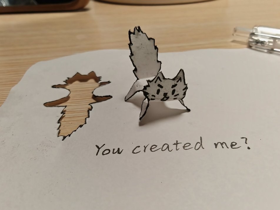A photograph of a handmade paper cut artwork featuring a simple, hand-drawn cat figure. The cat is cut out from white paper, with black ink details for its face (eyes, nose, mouth) and body outline, standing upright on the paper. To the left of the cat is the negative space cut-out hole in the paper, showing the wooden surface underneath. Below the cat, handwritten in black ink, is the text: 'You created me?'. The background includes a light wooden table surface, with a partial view of a silver stapler on the right and a blurred object (possibly a phone case) in the top left corner.