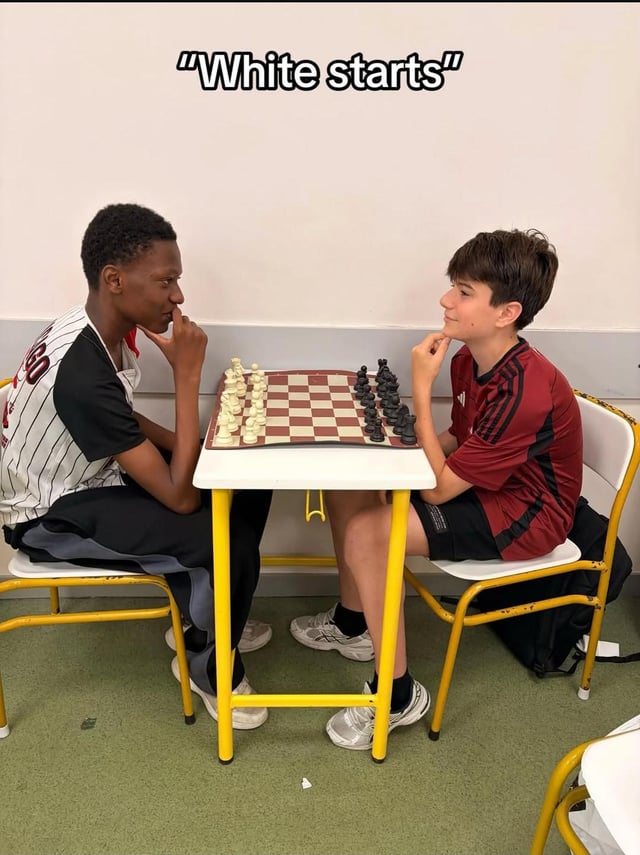 The image shows two teenage boys sitting opposite each other at a small table playing chess. A Black boy sits on the left with the full set of white chess pieces positioned in front of him, while a white boy sits on the right with the full set of black chess pieces in front of him. Text overlaid at the top of the image reads "White starts". The dark, ironic humor of the meme comes from a double entendre: in standard chess rules, the player using the white pieces always makes the first move, which would mean the Black boy on the left should start, but the phrase can also be read superficially as referencing the racial identity of the players, creating an awkward, edgy joke. Both boys look at each other with thoughtful, slightly amused expressions as if they both recognize the double meaning of the text.