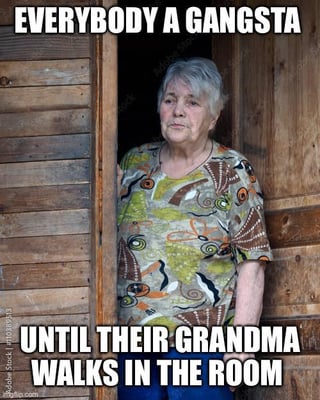 This meme features a stern-looking elderly woman (implied to be a grandma) standing in a rustic wooden doorway. Bold white text is overlaid at the top: **EVERYBODY A GANGSTA**, and at the bottom: **UNTIL THEIR GRANDMA WALKS IN THE ROOM**. The joke leans into the universal, relatable experience of people adopting a tough, 'gangsta' persona until confronted by a respected, authoritative family figure—here, a grandma—who immediately defuses that tough act. The grandma's unamused, serious expression reinforces the idea that she commands instant respect, making anyone abandon their tough demeanor quickly.