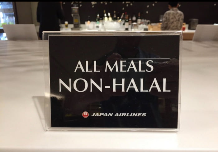 The image depicts a clear, rectangular sign placed on a white surface, likely in an airport lounge or airline service area. The sign has a black background with bold white text reading 'ALL MEALS NON-HALAL' at the top, and the 'JAPAN AIRLINES' logo (a red circle with a white stylized 'J' inside) followed by the airline name in white text at the bottom. In the background, there is a blurred bar or counter area with bottles and glasses visible, and two indistinct people standing, suggesting a public or service environment.