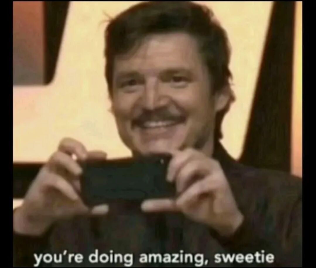 The image depicts a man with a mustache and short dark hair smiling warmly while holding a black smartphone in both hands, positioned in front of his face. The background is a blurred yellowish indoor setting, suggesting a stage or studio environment. At the bottom of the image, white text reads 'you're doing amazing, sweetie', conveying a supportive and encouraging message. The man's cheerful expression and posture indicate a positive, affirming demeanor, likely intended to uplift or praise someone.