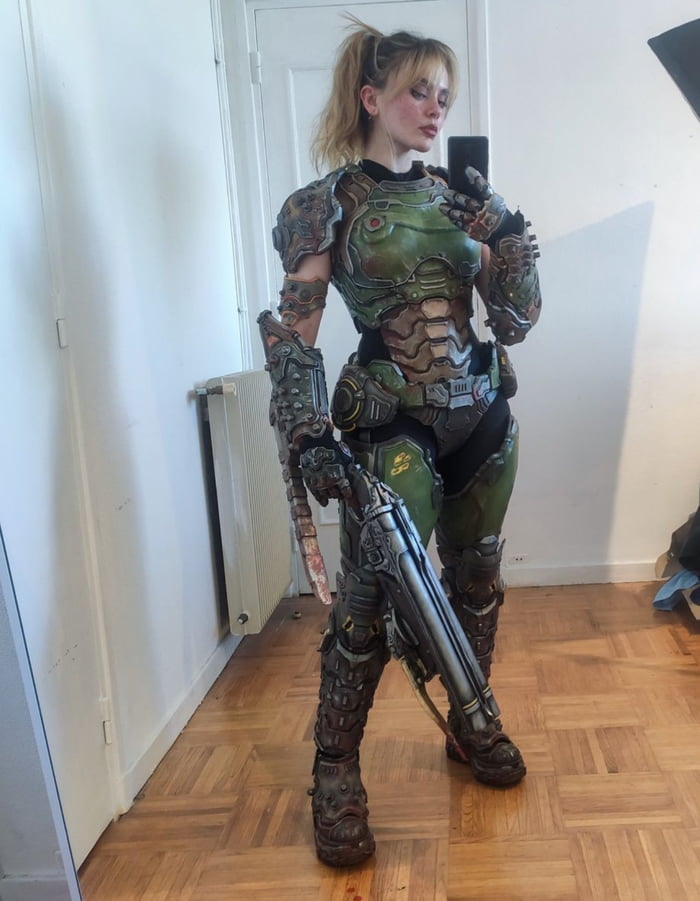 A person stands in a room with wooden flooring and white walls, wearing an elaborate green and brown armored costume that covers their entire body. The armor features intricate details including shoulder plates, chest armor, arm guards, and leg protection. The individual is holding a large, detailed futuristic weapon in their right hand while using their left hand to take a mirror selfie with a smartphone. The room has minimal furnishings including a white radiator visible along the wall.