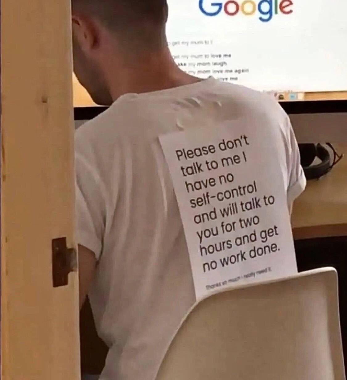 The image shows the back view of a man sitting at a desk facing a computer monitor displaying the Google search homepage. A printed paper sign is taped to the back of his plain white t-shirt, which reads: 'Please don't talk to me I have no self-control and will talk to you for two hours and get no work done.' The self-deprecating humor comes from the relatable admission that the wearer is easily distracted by casual conversation and will be unable to complete tasks if interrupted, so he is pre-emptively asking others not to speak to him to preserve his productivity. It resonates strongly with people working or studying in shared spaces who struggle to stay focused when approached by peers or coworkers.