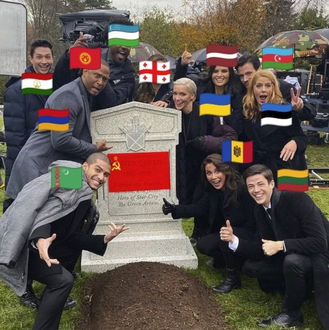 A meme image showing a group of smiling people (likely actors from the TV show 'Arrow') gathered around a tombstone labeled 'USSR 1922-1991' with a hammer and sickle symbol. The tombstone also includes text 'Area of Star City The Green Arrow,' referencing the fictional Star City from 'Arrow.' Overlaid on the individuals' faces are national flags of former Soviet republics (e.g., Ukraine, Lithuania, Latvia, Estonia, Armenia, etc.). The group displays celebratory gestures like thumbs up and peace signs, humorously marking the anniversary of the Soviet Union's collapse.