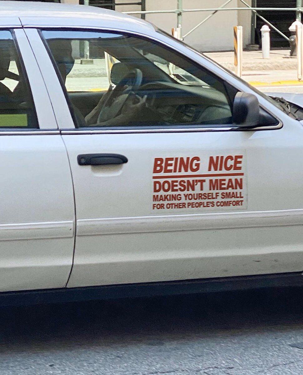 This is a candid street photograph showing the driver's side of a parked pale silver sedan. A red text decal is affixed to the front driver-side door, reading 'BEING NICE DOESN'T MEAN MAKING YOURSELF SMALL FOR OTHER PEOPLES COMFORT'. A person wearing a light-colored face mask is visible seated in the driver's seat inside the car. The background features a city sidewalk, metal scaffolding, and traffic bollards. The decal carries an uplifting, self-affirming message that encourages people to practice kindness without sacrificing their own boundaries, needs, or sense of self to make others comfortable.
