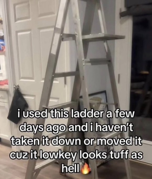 The image displays a silver folding A-frame step ladder left propped up in front of a white paneled interior door in a residential home setting, with part of a kitchen shelf visible to the left. Overlaid on the lower half of the image is white sans-serif text with a black outline, reading 'i used this ladder a few days ago and i haven't taken it down or moved it cuz it lowkey looks tuff as hell' followed by a small fire emoji. The meme humorously highlights the relatable, small lazy choice of leaving a household item out because you like its unexpected aesthetic.