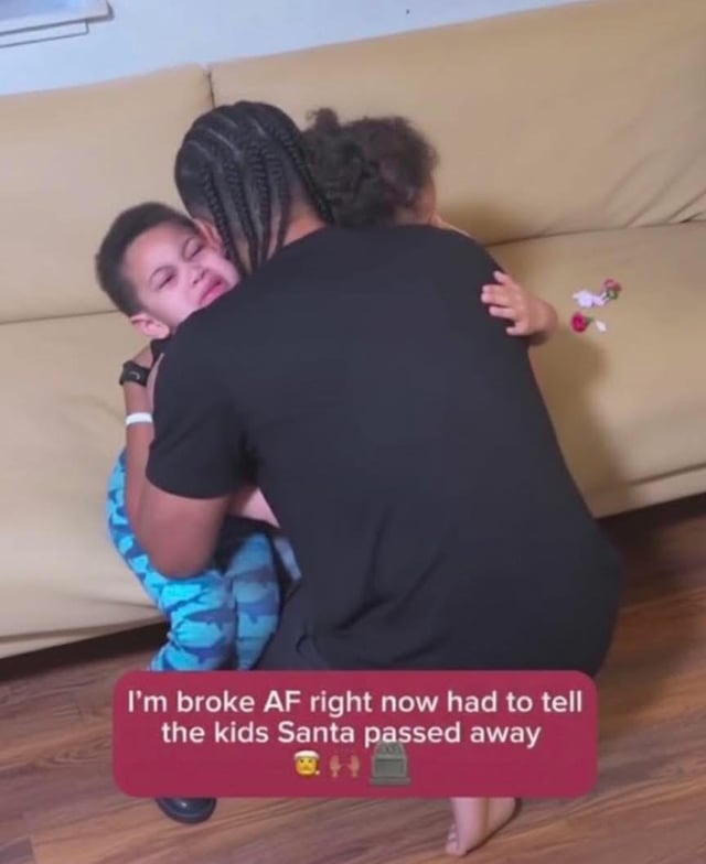 A photograph depicting a person with braided hair wearing a black shirt kneeling on a wooden floor, hugging two children on a beige couch. One child, dressed in blue camouflage-patterned pants, is visibly crying with eyes closed, while the other child rests their arm on the person's back. A red text overlay at the bottom of the image reads: 'I'm broke AF right now had to tell the kids Santa passed away' accompanied by a crying face emoji, folded hands emoji, and tombstone emoji. The scene blends a heartfelt moment of comfort with dark humor about financial hardship during the holiday season.