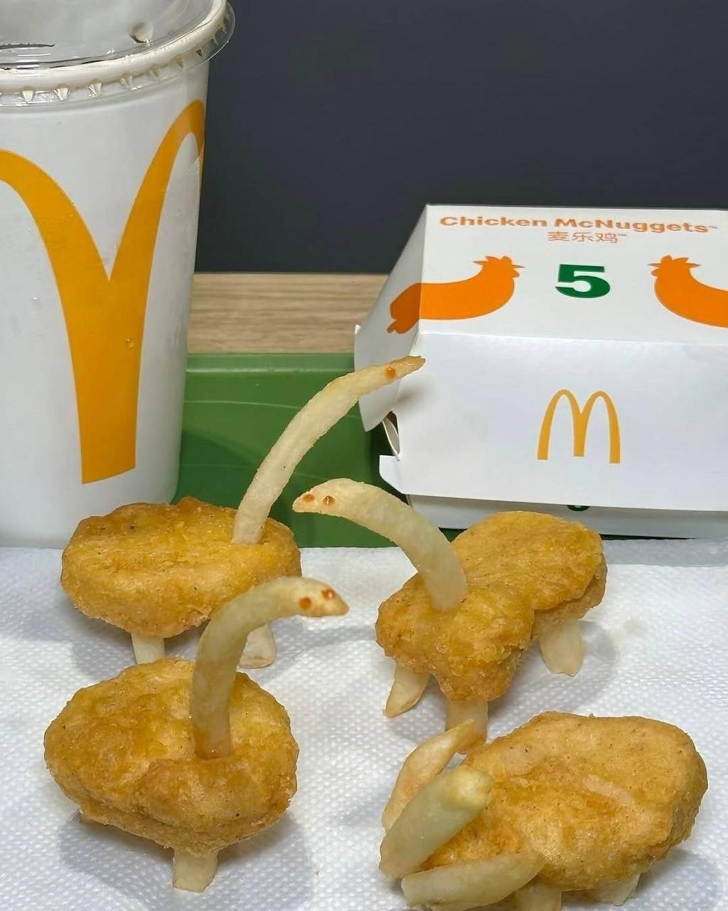 The image displays a McDonald's meal set on a tray, with a branded soda cup and a 5-piece Chicken McNuggets box visible in the background. In the foreground, the creator has arranged chicken nuggets as the bodies of small goose-like creatures, inserting French fries to serve as their necks (with tiny dots of sauce added as eyes) and legs, resulting in four cute, whimsical standing food figurines placed on a paper napkin. It is a lighthearted, playful piece of casual food art made entirely from standard McDonald's menu items.