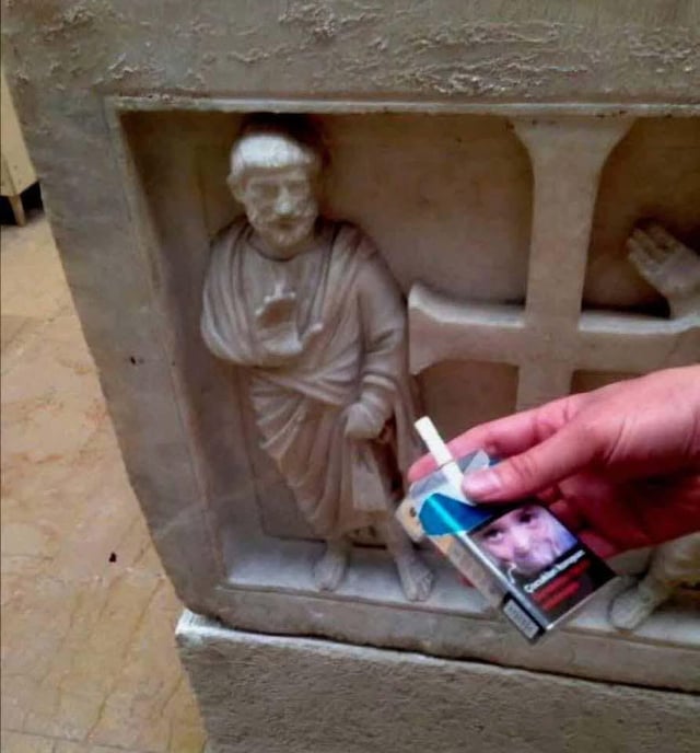 A photograph depicting a stone carving of a robed, bearded figure standing next to a cross-like structure. In the foreground, a human hand holds a pack of cigarettes with a cigarette protruding, positioned as if offering it to the carved figure. The humor stems from the absurd juxtaposition of a modern, casual action (offering cigarettes) directed at an ancient or religious stone carving, implying a polite refusal as suggested by the original title 'No thank you'.