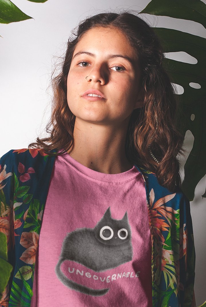 A person with long wavy hair wearing a pink t-shirt featuring a black cat with large round eyes and the word 'UNGOVERNABLE' written below the cat. They are also wearing a blue floral-patterned kimono with red, green, and white flowers. The background consists of large green leaves, creating a natural, tropical氛围. The person has a neutral expression and is looking slightly upwards.
