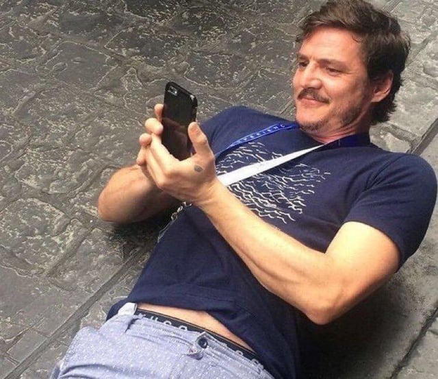 A man lying on a rough, textured stone floor, propped up on his side, holding a smartphone with both hands. He wears a dark blue t-shirt featuring a white abstract pattern, light blue shorts, and a lanyard around his neck. His expression is relaxed and subtly amused, as if he is reading something on the phone that gives him satisfaction. The setting suggests an outdoor or semi-outdoor area. This image is used as a meme to humorously depict the experience of re-reading one's own social media comment after it has accumulated a significant number of upvotes, capturing a relatable sense of pride or amusement.