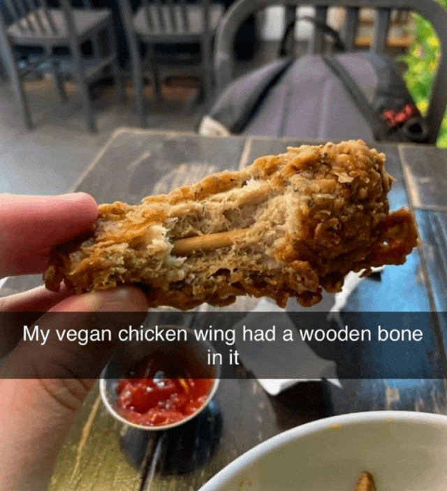 A photograph of a hand holding a vegan chicken wing, which has a visible wooden stick inserted to mimic a bone. The wing appears to have a breaded exterior and a textured interior, resembling traditional chicken. A text overlay at the bottom reads 'My vegan chicken wing had a wooden bone in it,' creating a humorous commentary on the realism of vegan meat substitutes, highlighting an unexpected and inedible wooden 'bone' as a quirky detail.