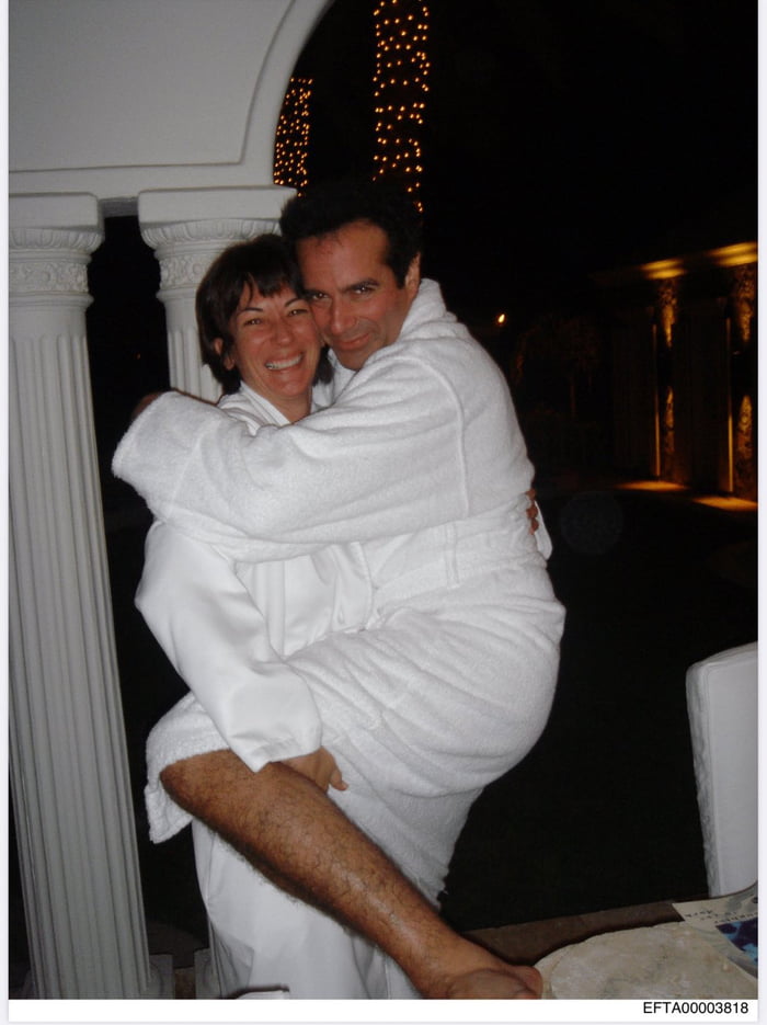 A nighttime photograph showing a man and woman embracing warmly outdoors. The man is lifting the woman in his arms, and both are wearing white robes. They are positioned in front of white decorative columns with intricate carvings. The background features string lights and warmly lit architectural elements, suggesting a resort or upscale outdoor setting. Both individuals have happy expressions and are smiling at the camera. The atmosphere appears romantic and joyful.