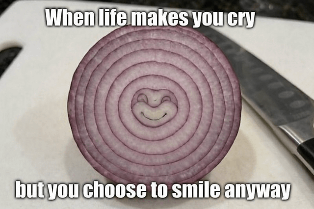 A meme featuring a sliced red onion placed on a white cutting board with a kitchen knife positioned beside it. The onion has a digitally added simple smiley face (two dots for eyes and a curved line for a mouth) in its concentric ring center. Above the onion, bold white text reads 'When life makes you cry,' and below it, additional bold white text states 'but you choose to smile anyway.' The humor stems from the pun on onions causing tears when cut, framing the onion as emotionally resilient by smiling through the metaphorical 'hardship.'