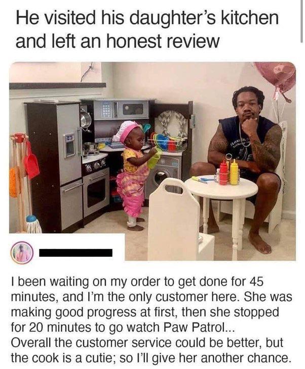 The meme includes a header text, a candid photograph, and a satirical customer review section. The header reads "He visited his daughter's kitchen and left an honest review". The photograph shows a toddler girl wearing a pink chef hat and apron, pretending to cook at a children's toy kitchen set, while her tattooed father sits at a tiny toy table across from her with a deadpan, unimpressed expression, resting his chin on his hand as he waits for his order, with small toy condiment bottles on the table. The review text below is written from the father's perspective, joking that he waited 45 minutes for his order as the only customer, his daughter initially made good progress but paused for 20 minutes to watch the children's show Paw Patrol, noting that customer service could be better but the cook is very cute so he will give her another chance. It is a lighthearted, endearing depiction of playful parent-child interaction.