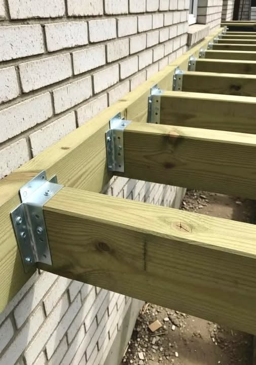 This image captures an active outdoor deck construction project. A series of pressure-treated wooden joists are being securely attached to a light-colored brick exterior wall using heavy-gauge metal joist hangers, each fastened with multiple screws. The perspective of the photo follows the line of joists as they recede into the background, emphasizing the uniform, repetitive installation pattern. The ground beneath the framing has scattered small construction debris, confirming that work is in progress. This is a standard, structurally sound method for building the foundational framework of an attached deck, ensuring the joists are properly anchored to the building for long-term stability and support.