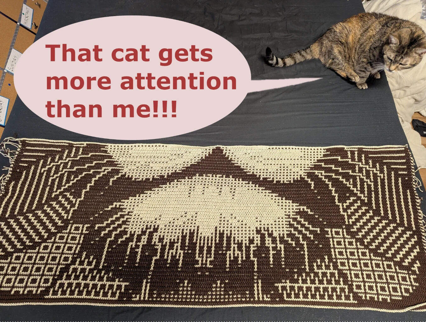 The image features a real tabby cat with brown and black fur sitting on a dark fabric surface. To the left of the cat is a large, handmade knitted piece displaying an intricate geometric pattern in cream and dark brown, resembling abstract mountain or wave designs. A prominent pink speech bubble with bold red text overlays the upper left portion of the image, reading: 'That cat gets more attention than me!!!' The cat appears calm, looking downward with its tail curled beside it, creating a humorous contrast between the cat's nonchalant demeanor and the unseen speaker's playful complaint about the cat receiving more attention than their crafted project.