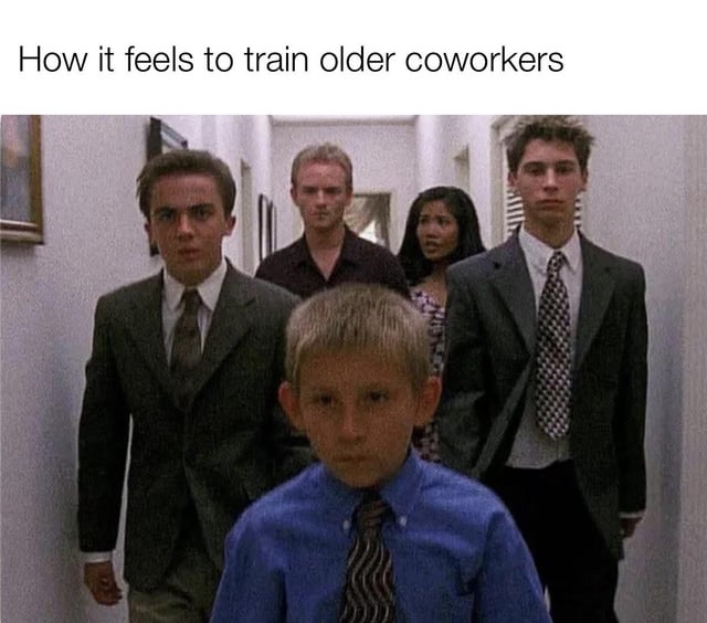The meme features a screenshot from a movie hallway scene. In the foreground, a young boy wearing a blue shirt and tie walks forward with a serious expression. Behind him, four older individuals (three men and one woman) dressed in formal suits follow closely, also with neutral to serious expressions. The text at the top reads 'How it feels to train older coworkers,' humorously reversing the typical dynamic by portraying the younger person as the leader/trainer and the older individuals as the ones being trained, highlighting the relatable workplace scenario of younger employees training older colleagues.