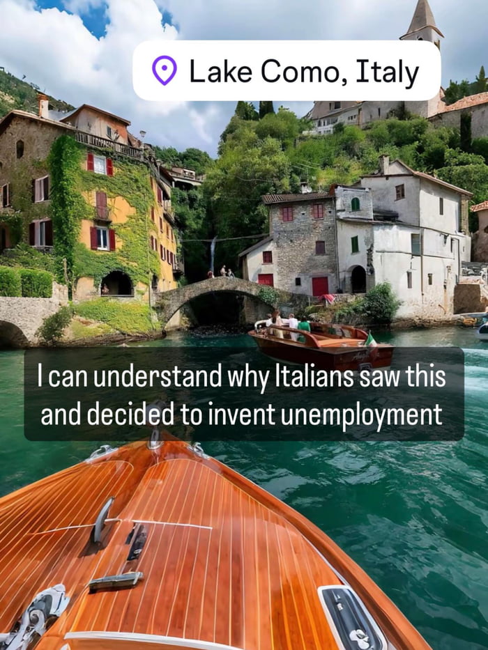 This is a first-person travel photograph taken from the bow of a polished wooden boat floating on bright turquoise water at Lake Como, Italy. The scenic background features picturesque historic lakeside buildings partially covered in ivy, a small stone arch bridge, a cascading waterfall, lush green forested hills, and a stone church spire, with another wooden leisure boat carrying passengers visible on the water. A humorous text overlay makes a lighthearted playful joke that leverages the common stereotype of Italian work culture prioritizing leisure and work-life balance, implying the idyllic scenery is so beautiful it would make people choose not to work to enjoy their surroundings.