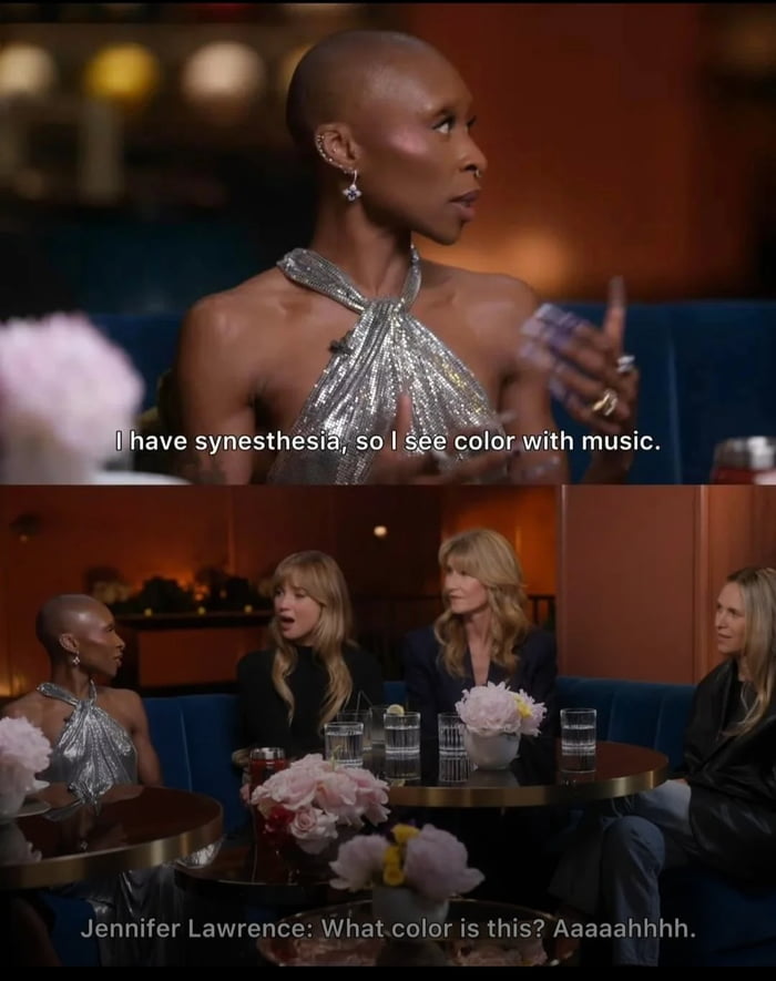A two-panel screenshot from a talk show or interview setting. The top panel shows a person with a shaved head wearing a shiny silver halter dress, gesturing while speaking; text overlay reads, 'I have synesthesia, so I see color with music.' The bottom panel shows the same person seated on a blue couch with three other women. One of the women, identified by text as Jennifer Lawrence, reacts with an excited/shocked expression, saying, 'What color is this? Aaaaaahhhh.' The setting includes small tables with water glasses and pink flower arrangements, suggesting a casual, conversational atmosphere.