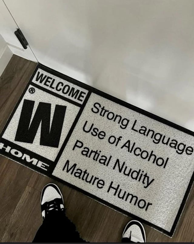 The image displays a novelty welcome doormat positioned on a wooden floor adjacent to a white interior door. At the bottom of the frame, the feet of a person wearing black-and-white low-top sneakers are visible, standing just in front of the mat. The doormat uses a direct parody of the Entertainment Software Rating Board (ESRB) content rating label format: on the left side, a large, bold 'W' (styled after ESRB's rating icons) sits between the text 'WELCOME' at the top and 'HOME' at the bottom. On the right side, it lists four content descriptors typically found on mature-rated video game labels: 'Strong Language', 'Use of Alcohol', 'Partial Nudity', and 'Mature Humor'. The core joke is framing a private home as if it were a mature-rated media product, playfully warning visitors about the casual, unfiltered, adult-oriented environment they are entering. This relatable humor resonates with adults who have a laid-back, informal household dynamic.