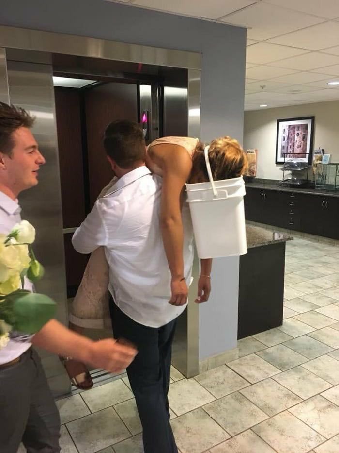 This is a real-life viral photograph taken in a tiled commercial lobby next to an open elevator. A man presumed to be a groom wears a white dress shirt and carries a woman presumed to be his bride, who is wearing a formal light-colored dress, over his shoulder. The woman is completely limp and heavily intoxicated, with her entire head positioned inside a white plastic bucket hanging off the back of the man to catch vomit if she gets sick. A second man holding a bouquet of white roses walks nearby, smiling at the absurd scene. The image functions as a wedding fail meme, playing on the subversion of expectations for a romantic, elegant post-wedding experience, with the practical but ridiculous setup of the bucket holding the bride's head becoming the core visual joke.