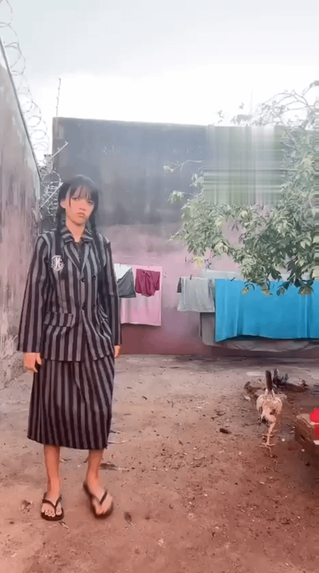 A meme image showing a young woman standing in a rural courtyard setting. She is wearing a black and white striped outfit that resembles a school uniform or pajamas, paired with flip-flops. The woman has a neutral expression on her face and is standing still with her arms at her sides. Behind her, there are clothes hanging on a line to dry, including pink, gray, and blue items. A chicken is walking on the dirt ground near her feet. The setting features a dirt ground, concrete walls, and some vegetation. The original title suggests a comparison between Wednesday (marked with ❌) and Thursday (marked with ✅), implying the woman's mood or situation improves from one day to the next.