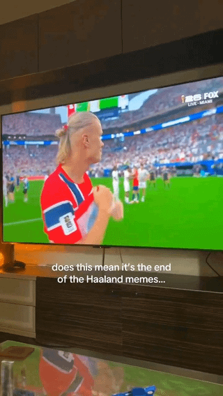 This image is a personal photograph of a television screen in a home setting, displaying a live FOX Sports broadcast of a football (soccer) match. The broadcast features professional player Erling Haaland wearing a Norway national team jersey, standing on the pitch with his hands clasped while looking off to the side. White overlaid text at the bottom of the photo reads 'does this mean it's the end of the Haaland memes...', referencing the large volume of popular internet memes made featuring Haaland, joking that the end of the football playing season will mean no new source content for these memes. The television sits on a dark wooden TV stand, with a reflection of the screen visible on a glass table below the stand.